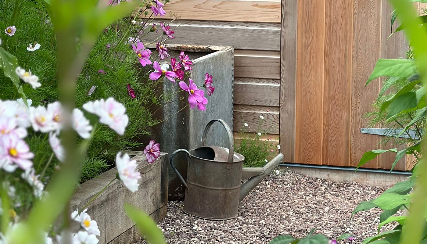 Rustic watering can sat outside of a garden shed surrounded by beautiful flowers