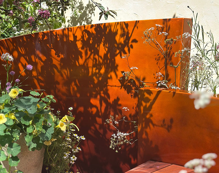 Orange garden metal panels with flower shadows surrounded by potted and wildflowers in sunlight.