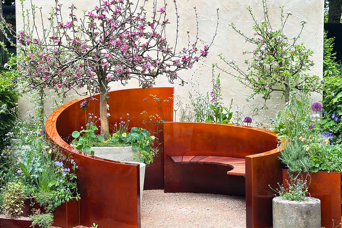 Curved rust-colored metal garden bench surrounded by flowering plants and small trees in planters.